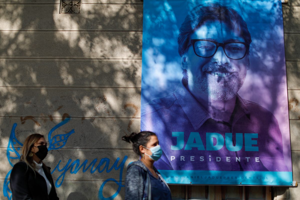 La fotografía muestra un pendón con el rostro de Jadue y dos personas caminando en la calle. 
