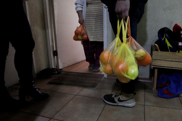 La imagen muestra a tres personas, dos de ellas con bolsas de comida en las manos, en el pasillo de un edificio. 